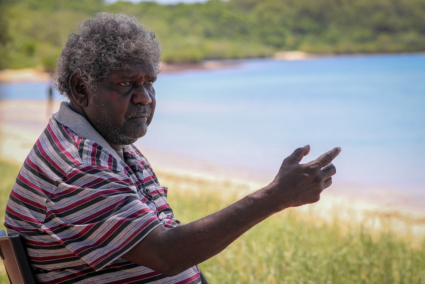 A man wearing a T-shirt pointing and talking, while sitting on the beach.