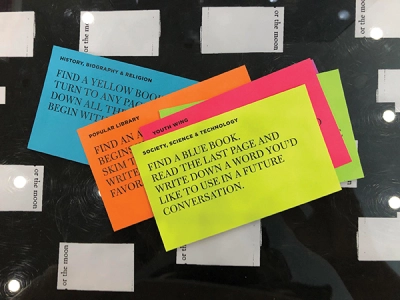 Multicolored index cards printed with instructions for activities to perform in a library lie on a black surface