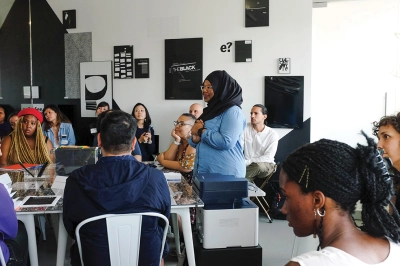 A dark-skinned woman wearing a black headscarf stands in the middle a room of seated people with thoughtful expressions. Everyone is looking at and listening to someone off-camera