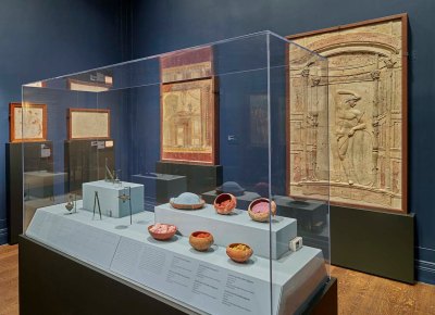 An installation view of a gallery shows a vitrine in which small bowls of pigment are displayed alongside metal implements. On the walls hang frescoes.