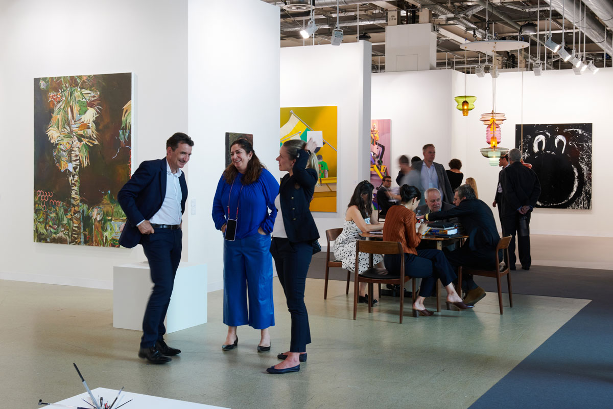 A group of three people stand near the aisles of an art fair with people at a table seated behind them. 