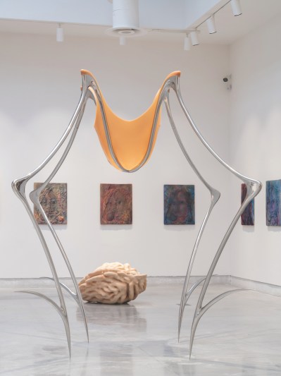 A gallery install view shows a sculpture in the foreground with a metal armature resembling a four-legged spider with silicone stretched over the top. In the background is what looks like a giant peach pit and a series of paintings hung on the wall.