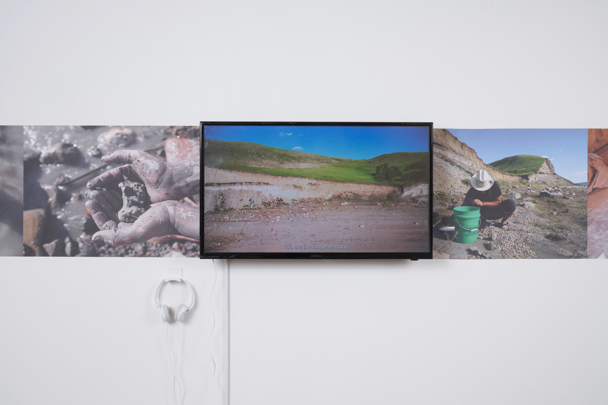 An installation view shows a TV playing a video depicting a landscape with grass and hills, surrounded by prints on the walls of hands holding clay and a person scooping something from the ground into a bucket.